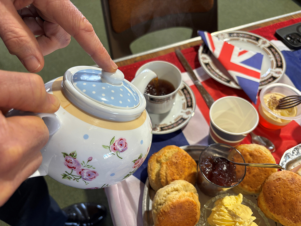 Tea being poured from a china teapot into a teacup, which is placed next to a plate of scones and jam in one of the tables